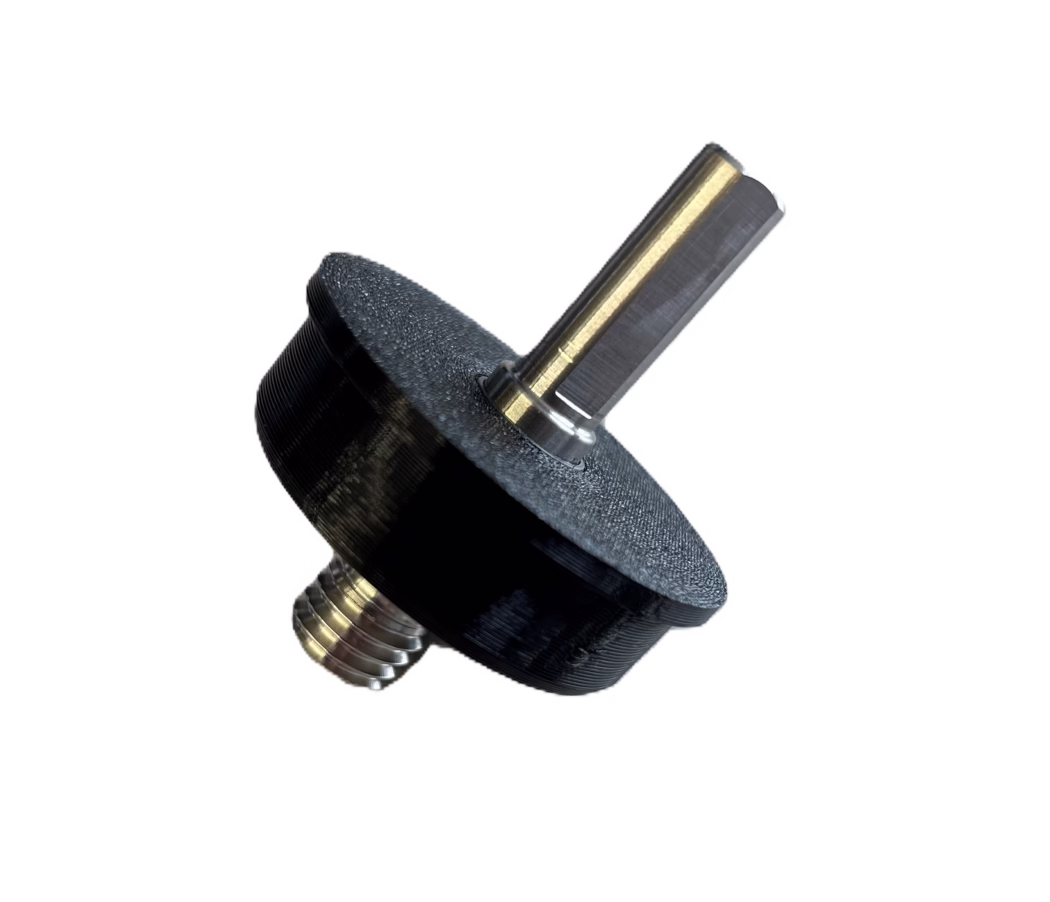 PALLMANN_product image_adapter cordless screwdriver Närbild av en liten mekanisk komponent: en sexkantig metallaxel med mässingsfärgad beläggning infogad i en svart plast- eller 3D-printad ratt eller distans. Metallaxeln har en polerad plan yta och synliga svarvspår; en gängad tapp med koncentriska gängor syns vid basen. Den svarta delen har en texturerad, lagerstrukturtypisk yta för FDM 3D-printing. Objektet är fotograferat diagonalt mot en ren vit bakgrund.